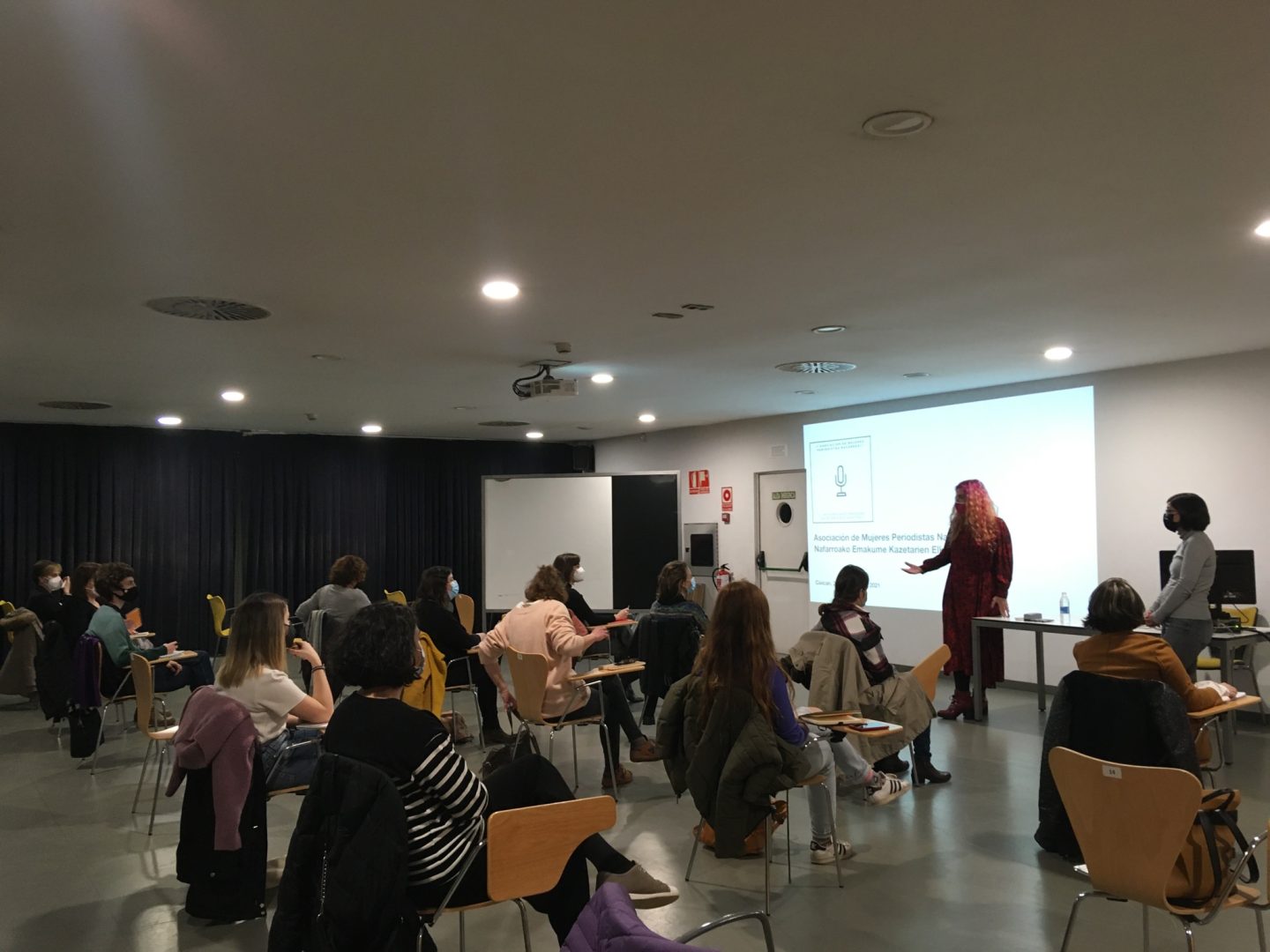 Presentación de la Asociación de Mujeres Periodistas. María Vallejo y Diana González presentan, ante una pantalla, los principios de la asociación con una vista general de la sala con varias personas sentadas.